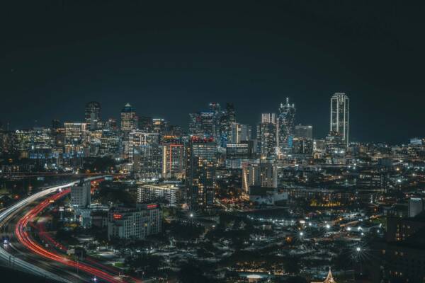 Car tail lights trailing in the night under the Dallas skyline.